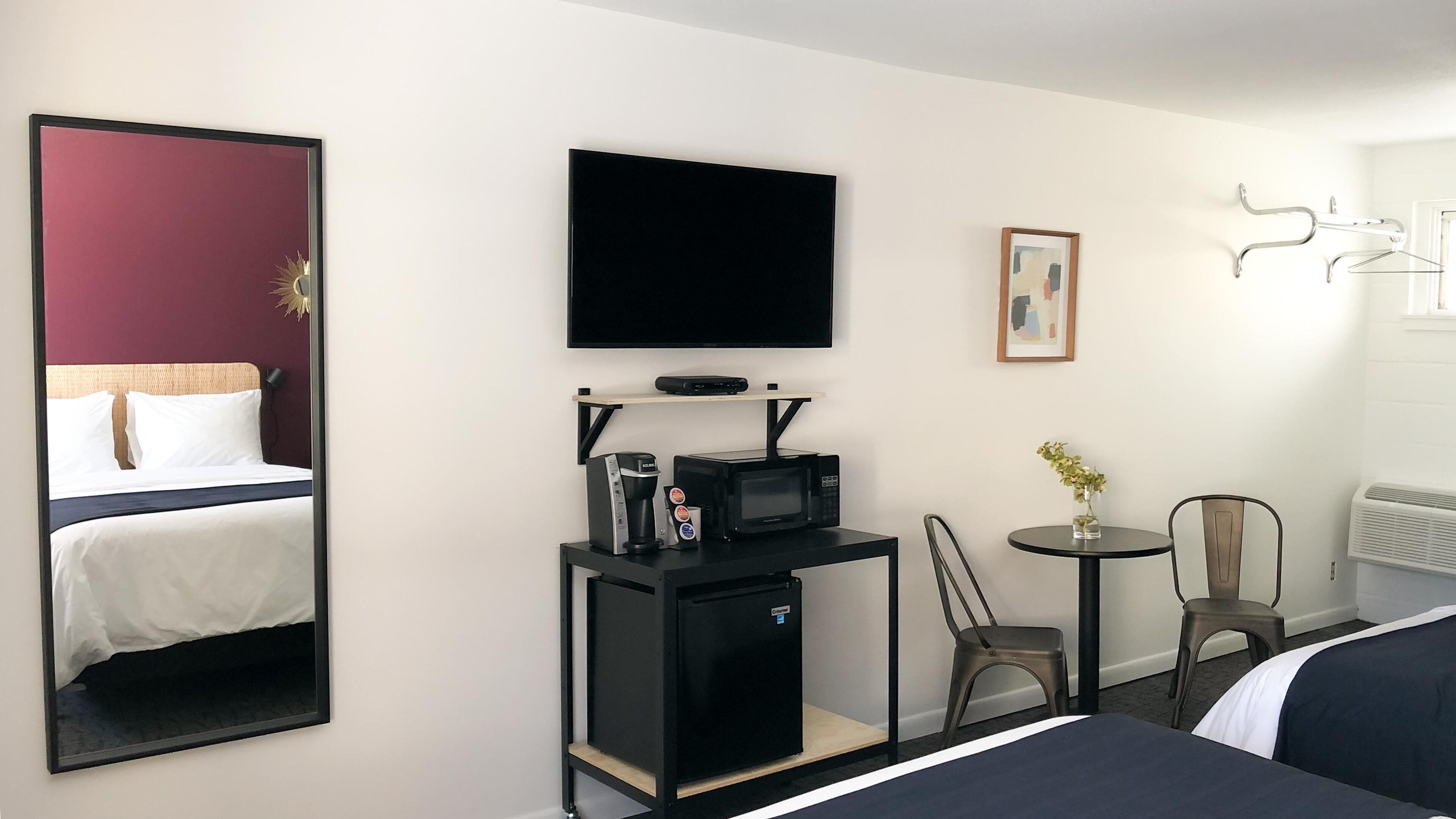 Habitación de hotel con cama, televisor, refrigerador pequeño, microondas, cafetera, silla, mesa con un florero y calefactor.