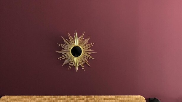 A bed with white bedding, a headboard with a woven texture, and a wall-mounted sunburst mirror.