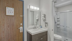 A bathroom with a wooden door, a sink with a mirror, a towel rack, and a glass shower enclosure.