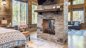 A log cabin interior with a stone fireplace, large windows, and a cozy seating area. - Somewhere With You -Features a hot tub & fire pit; accommodates 2 to 4 guests.   (Broken Bow)