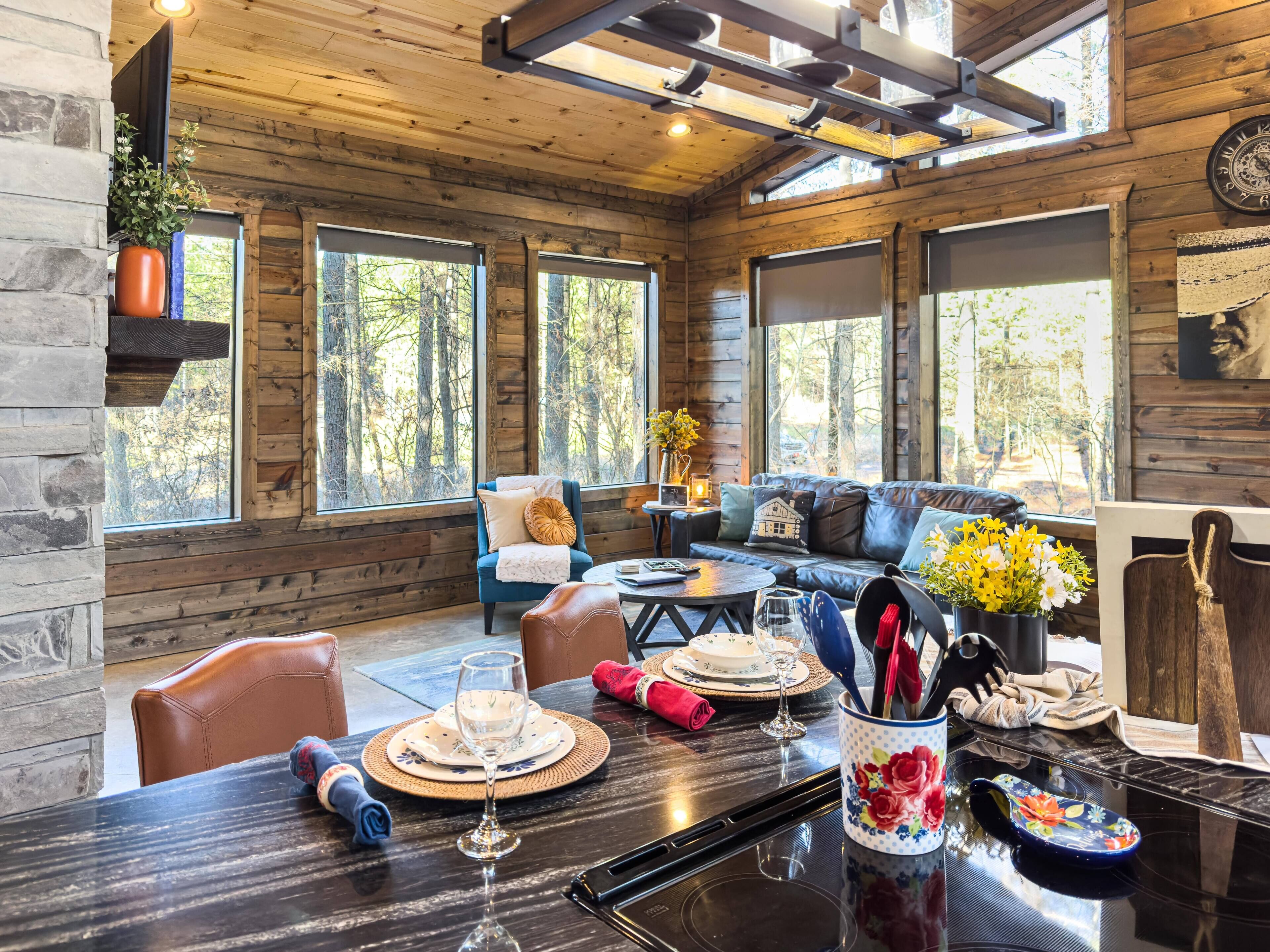 A cozy cabin interior with a dining area, a sofa, and large windows offering a view of the forest.