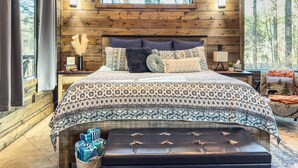 Ein Schlafzimmer in einer Blockhütte mit einem großen Bett, einer Bank und Blick auf Bäume durch das Fenster.