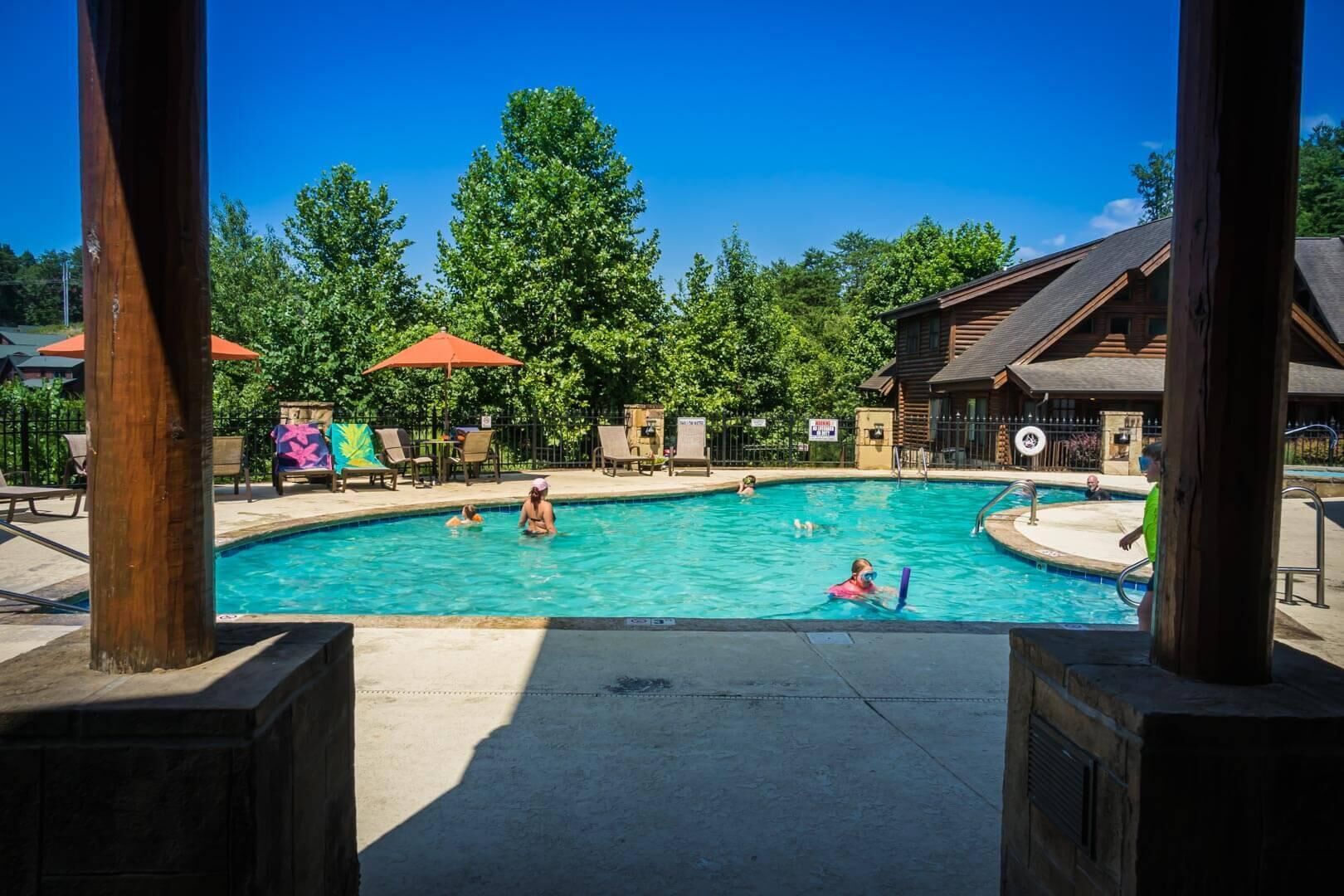 A pool area with multiple people swimming and lounging, surrounded by wooden cabins and trees.