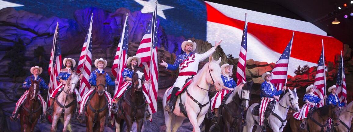Um palco com artistas trajados como cowboys montados em cavalos, segurando bandeiras americanas.