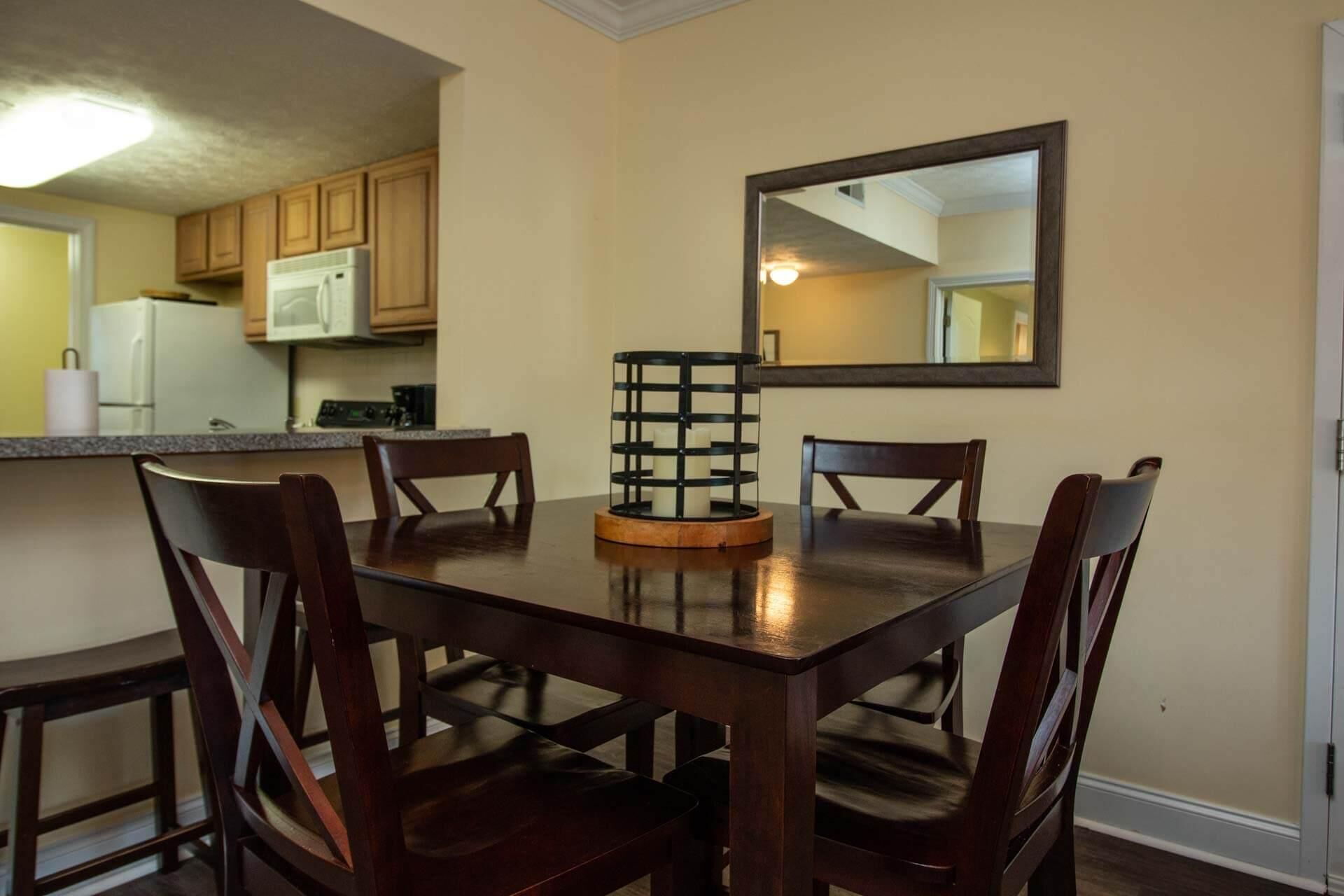 A dining area with a wooden table and four chairs, a candle holder, and a kitchen in the background.