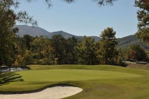 Um campo de golfe com um bunker de areia, árvores e montanhas ao fundo.
