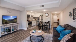 A modern living room with a TV, a coffee table, and a sofa with blue cushions. There is a kitchen in the background with white cabinets and a dining area with a table and chairs. - 2501 Golf View Towers Almost Home (Pigeon Forge)