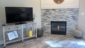 A living room with a flat-screen TV on a wooden console, a stone-patterned fireplace, and a round wall clock. - 1404 Golf View Towers Smoky Mtn Bearadise (Pigeon Forge)