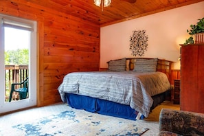 A wooden cabin bedroom with a bed, a decorative wall piece, a window, and a view of the outdoors.