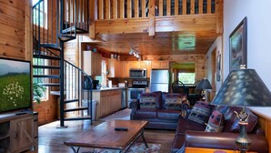 A cozy cabin interior with a wooden staircase, a kitchen area, a living room with a sofa and a coffee table, and a television displaying a nature scene. - 839a Golf View Cabins Rockin' Robin (Pigeon Forge)
