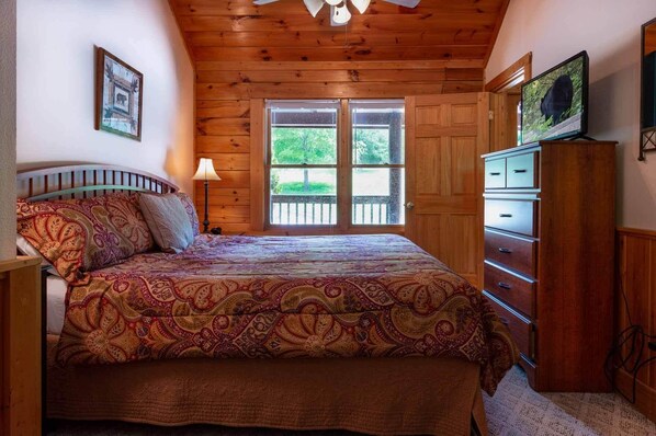 A bedroom with a wooden ceiling, a bed with a patterned bedspread, a nightstand, a lamp, a dresser, and a television on the wall.