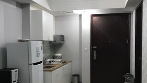 A compact kitchen with white cabinets, a refrigerator, a microwave, and a small countertop. There is a door leading to another room and a trash bin near the door.