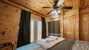 Une chambre aux murs lambrissés, avec un lit, un ventilateur de plafond et une vue sur l’extérieur.