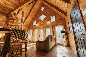 Interior d'una cabana de fusta amb un llit en altell, un sofà, terres de fusta i grans finestres.