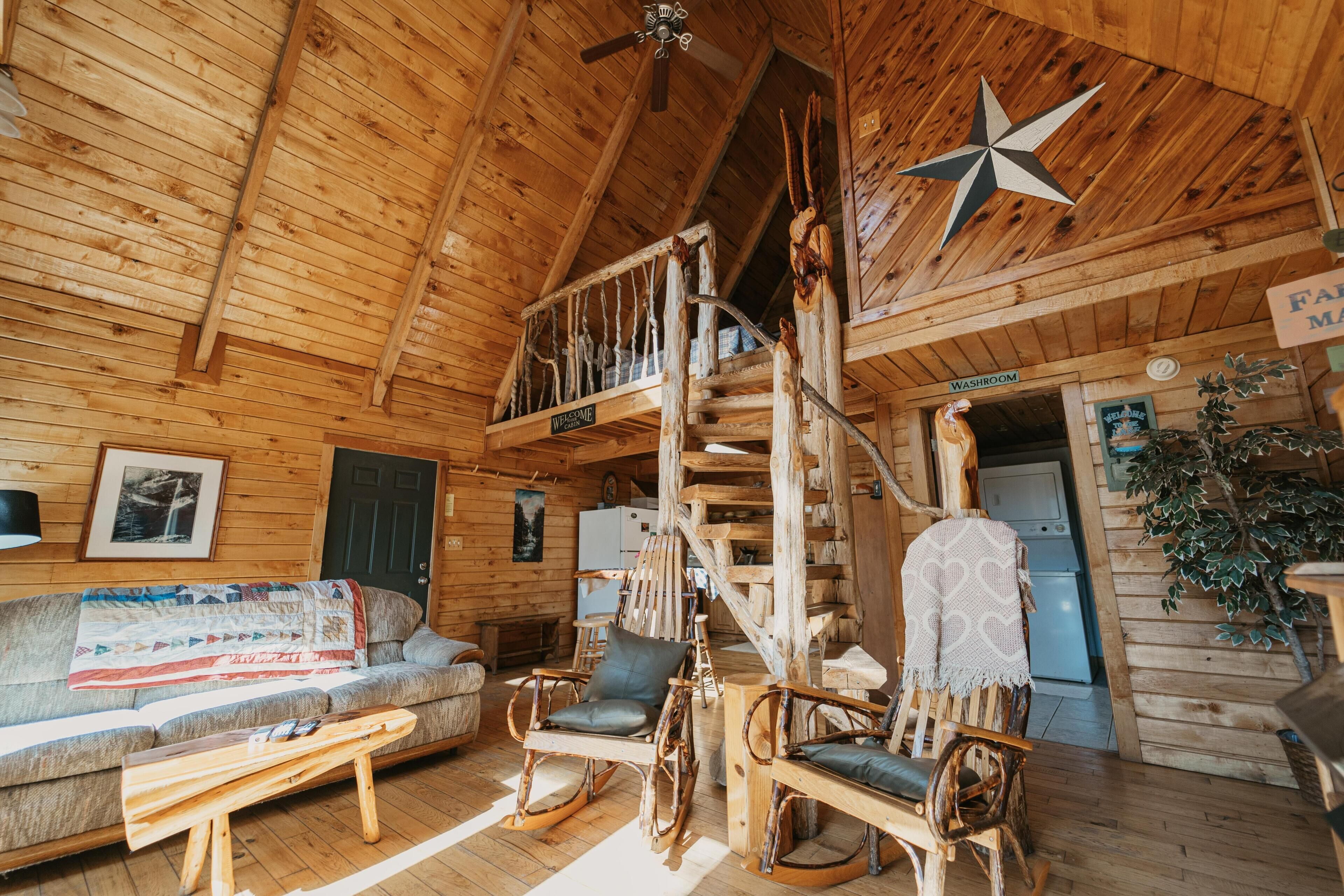 L’intérieur d’un chalet en bois, avec un canapé, des fauteuils à bascule, un escalier et un panneau indiquant les toilettes.