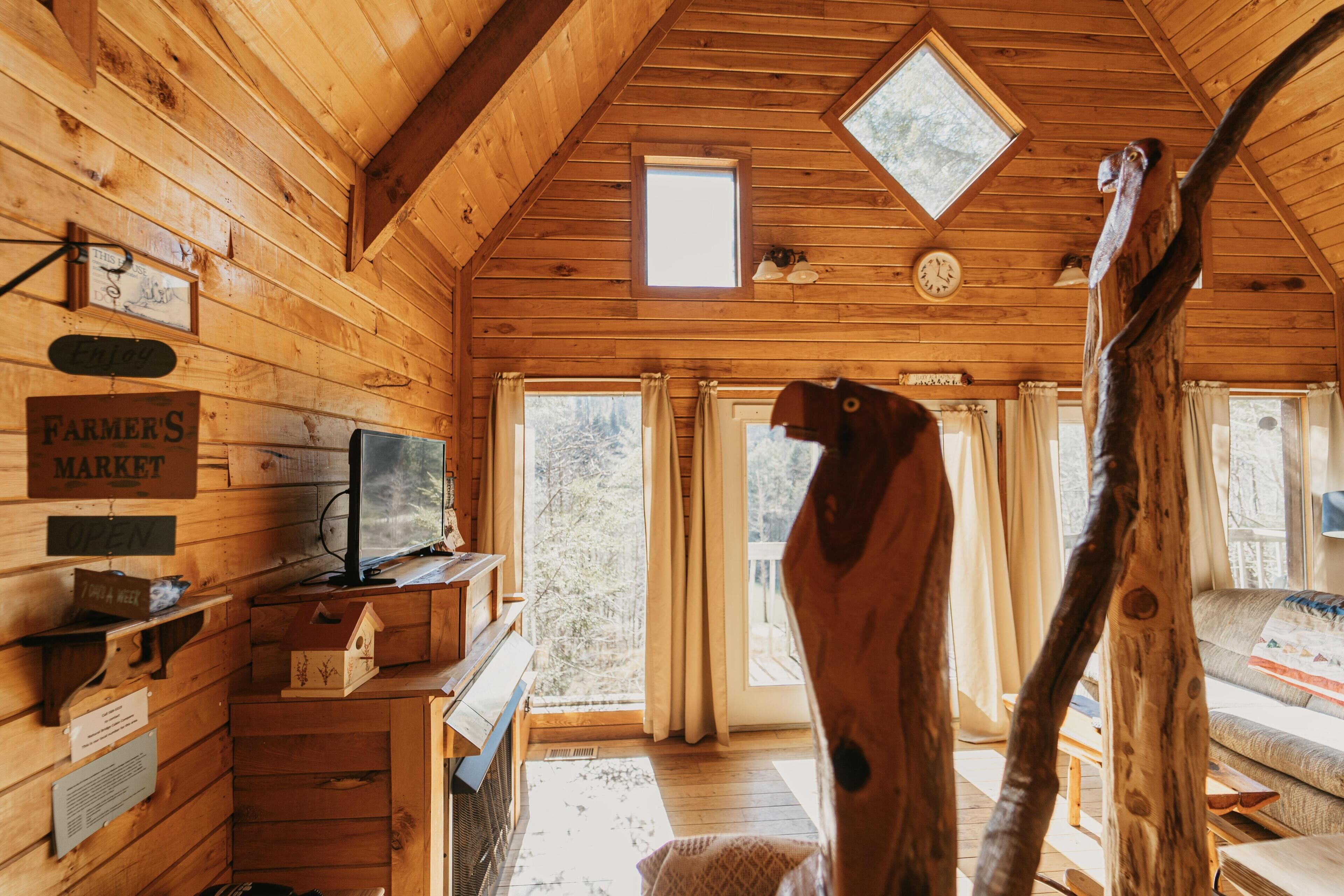 Interior de una cabaña de madera con cama en altillo, una silla de madera, televisor y ventana con cortinas.