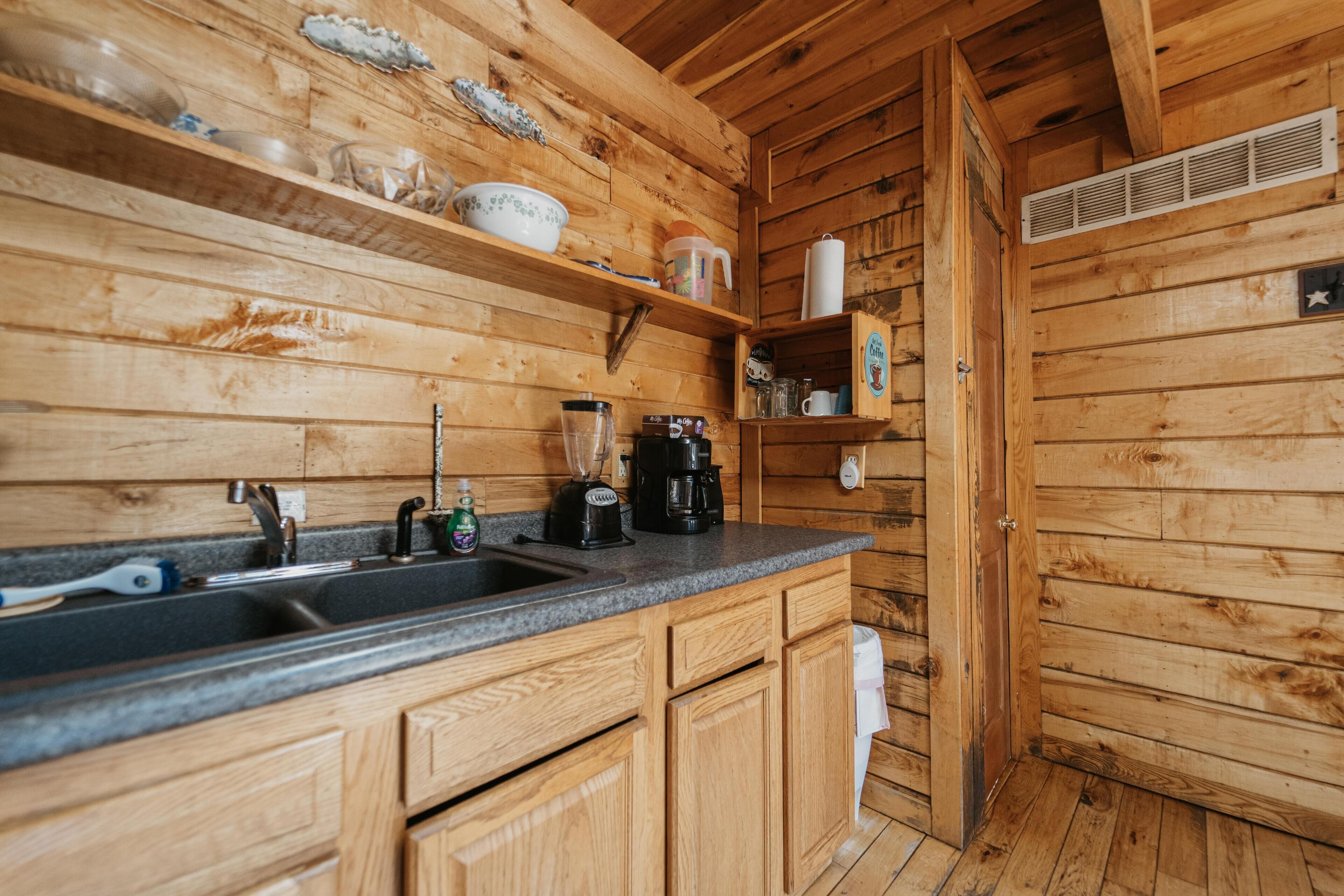 A wooden cabin kitchen with a granite countertop, wooden cabinets, a sink, a coffee maker, and a shelf with dishes and a mug.