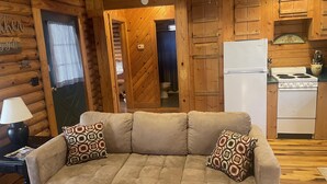 A cozy log cabin interior with a beige sofa, patterned pillows, a small table with a lamp, and a kitchen area with a stove and refrigerator.