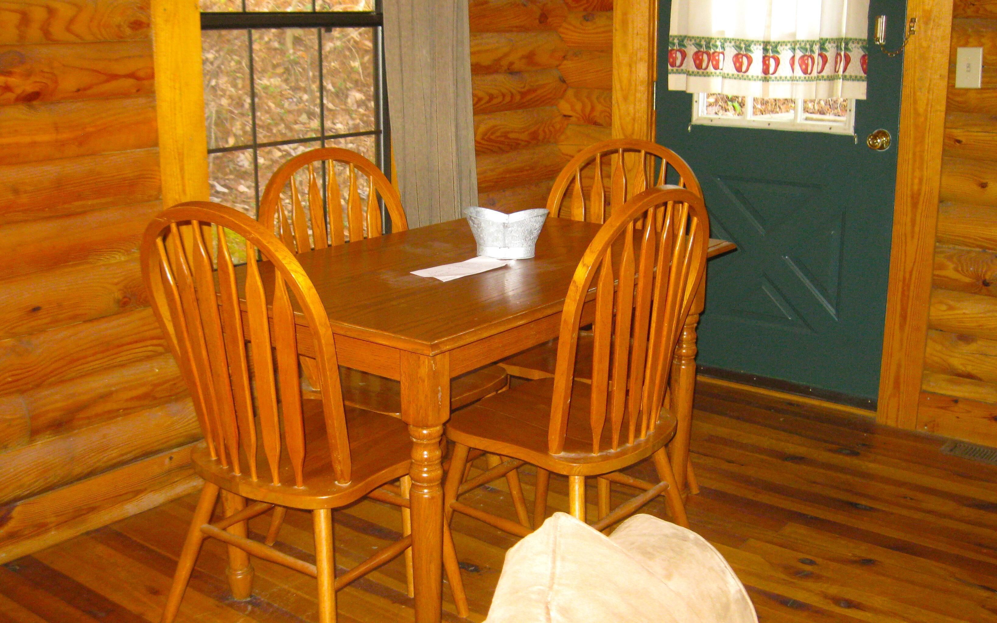 A wooden dining table with four chairs, a window with a view of trees, and a door with a fruit-themed curtain.