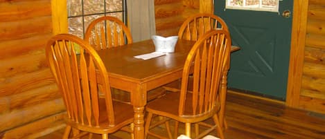 A wooden dining table with four chairs, a window with a view of trees, and a door with a fruit-themed curtain.