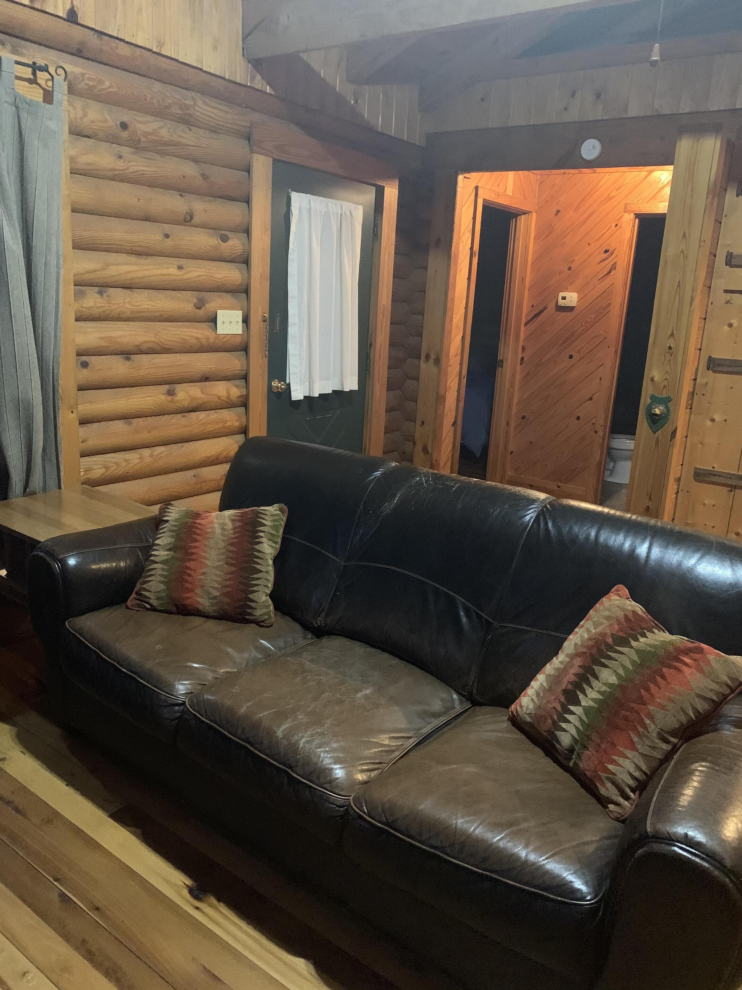 Interior de una cabaña de troncos con un sofá de cuero y dos almohadones, paredes de madera y una puerta que da a otra habitación.
