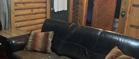 A log cabin interior with a leather sofa and two pillows, wooden walls, and a door leading to another room.