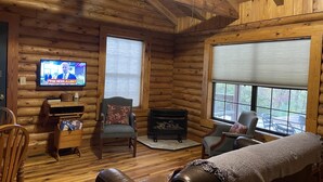 A log cabin interior with a television displaying a news program, a fireplace, and a cozy seating area with a couch and chairs. - Cabin close to Natural Bridge State Park with hot tub and internet (Slade)