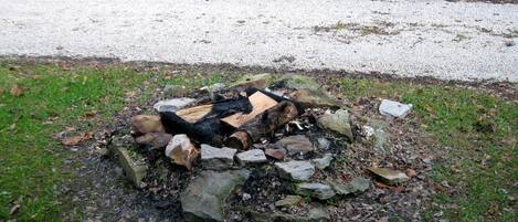Una fogata rodeada de rocas y troncos en un área con césped, con un sendero de grava al fondo.
