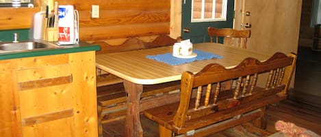 A wooden cabin interior with a kitchen area, a dining table, and a bench.