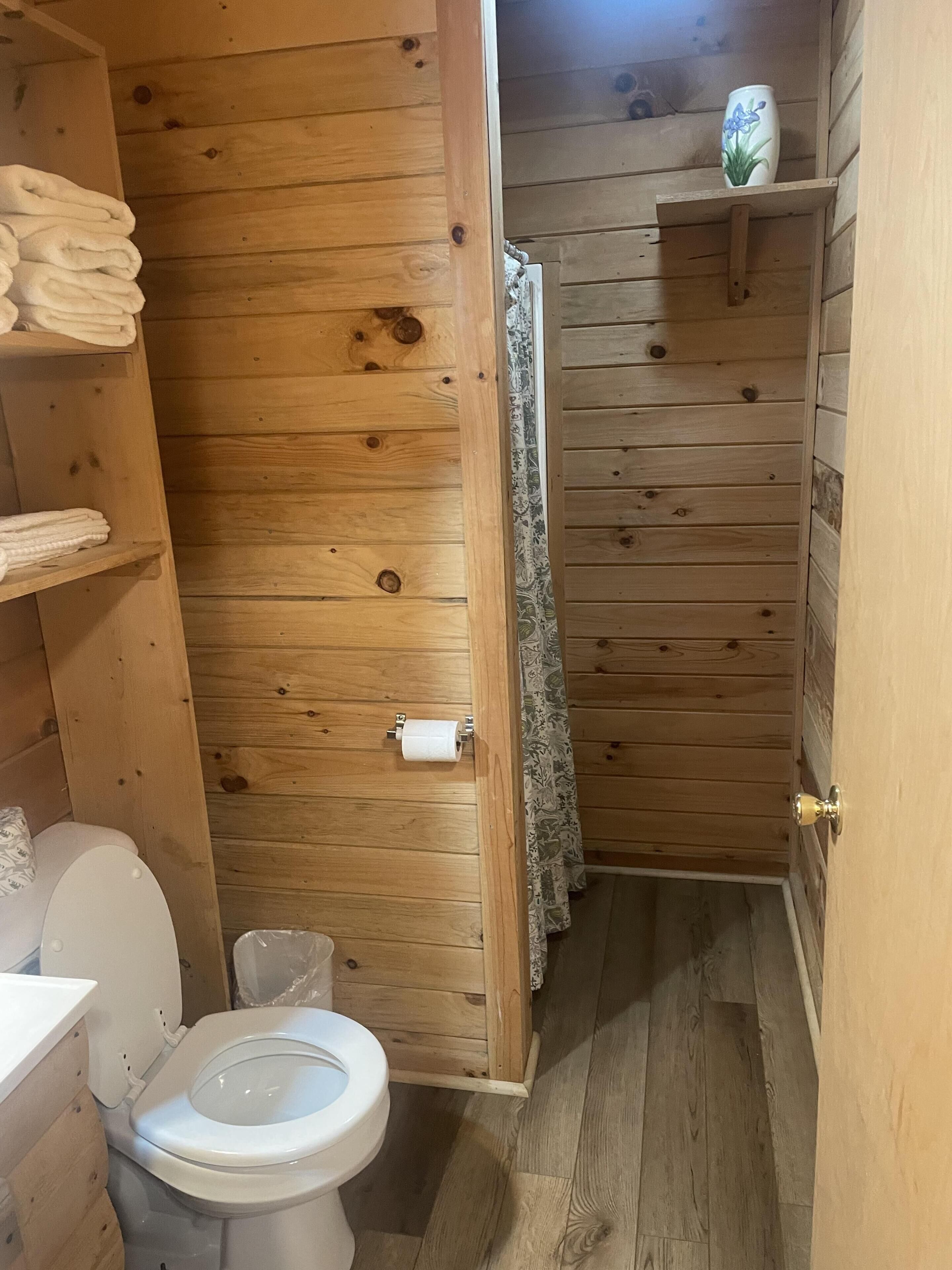 A wooden bathroom with a toilet, a shelf with towels, and a shower area.