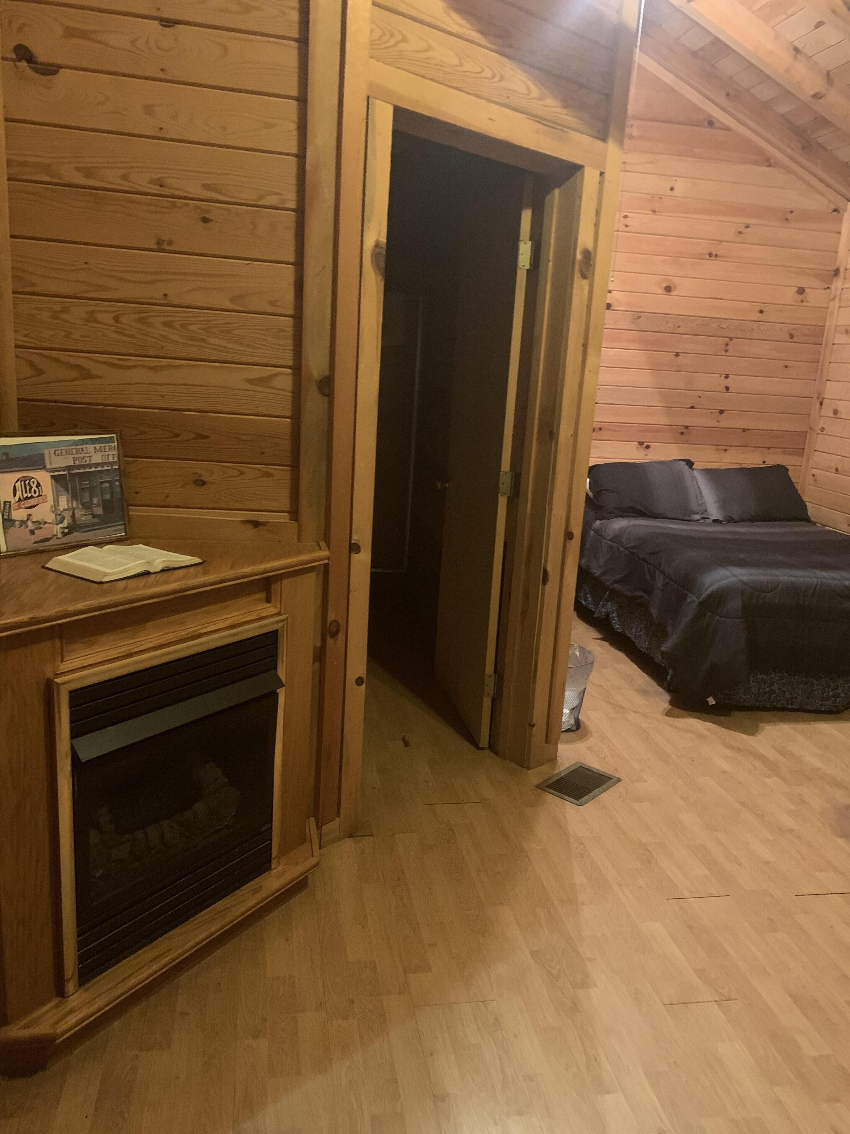 A wooden cabin room with a bed, a fireplace, and a door leading to another room.