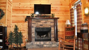 A cozy log cabin interior with a stone fireplace, a television mounted above it, and a plaid-patterned chair. - Spacious cabin with Hot Tub and wood burning fireplace and wi-fi (Campton)