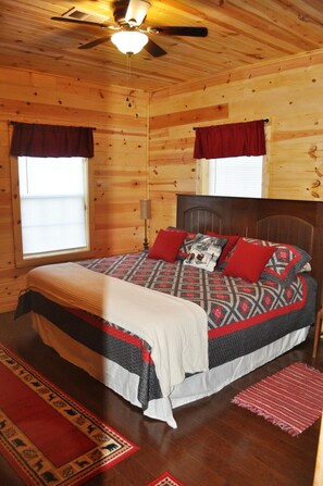 Un dormitorio de cabaña de madera con una cama, un ventilador de techo y dos ventanas con cortinas.