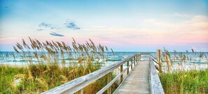 A wooden boardwalk leading to a beach with tall grasses and a clear sky.