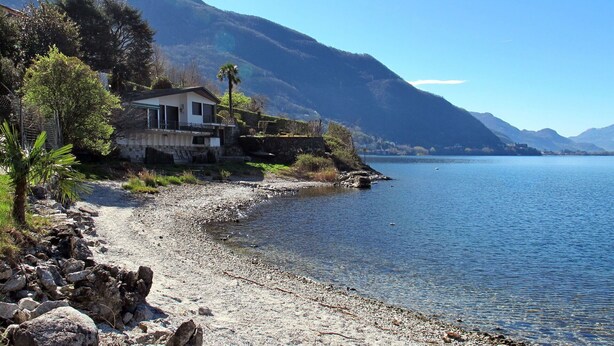 Una casa junto a un lago, con orilla rocosa y rodeada de montañas.
