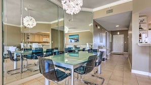 A modern dining area with a glass table and chairs, a kitchen in the background, and a large mirror reflecting the space. - Gulf Views & Pool Access: Coral Reef Resort Condo (Panama City Beach)