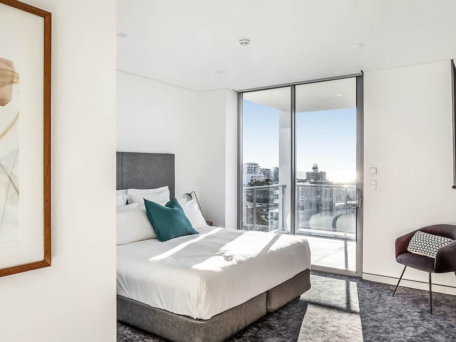 Quarto de hotel moderno com uma cama grande, vista da cidade através de uma porta de vidro deslizante e uma obra de arte emoldurada na parede.