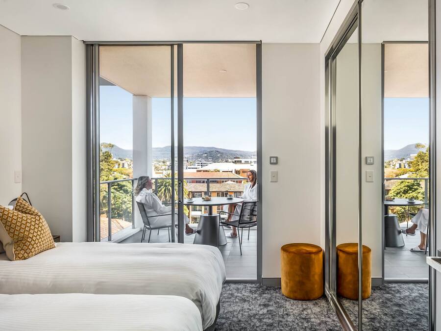 A modern hotel room with a large bed, a balcony with a view of the city, and two round wooden stools.