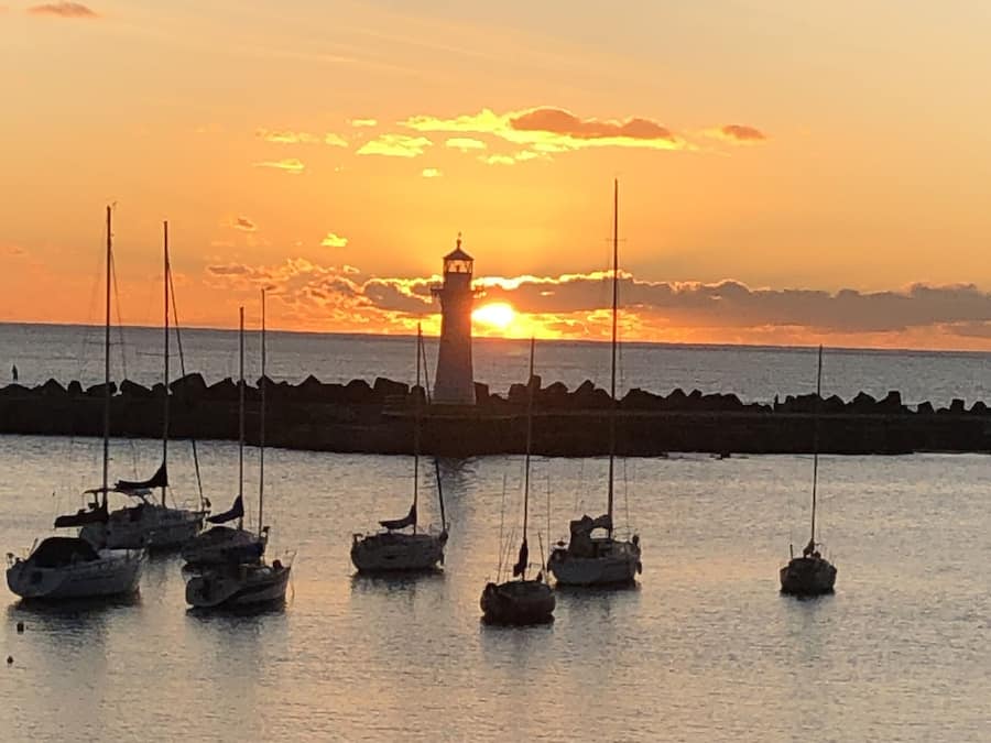Um porto ao pôr do sol com vários veleiros ancorados perto de um farol.