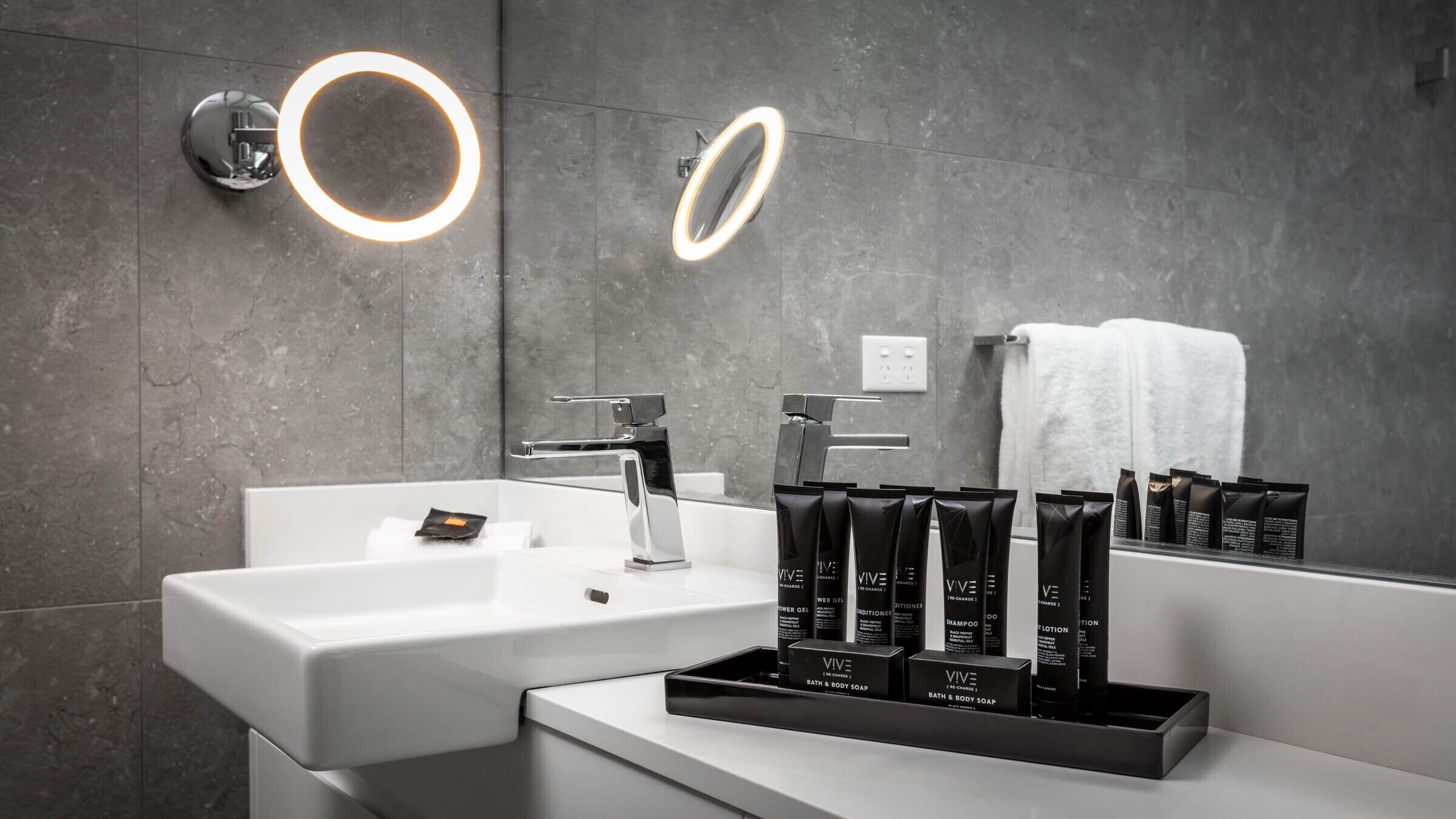 Ein modernes Badezimmer mit einem weißen Waschbecken, zwei beleuchteten Spiegeln und einem Waschtisch mit Pflegeprodukten.