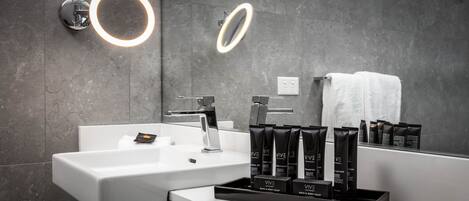 Ein modernes Badezimmer mit einem weißen Waschbecken, zwei beleuchteten Spiegeln und einem Waschtisch mit Pflegeprodukten.