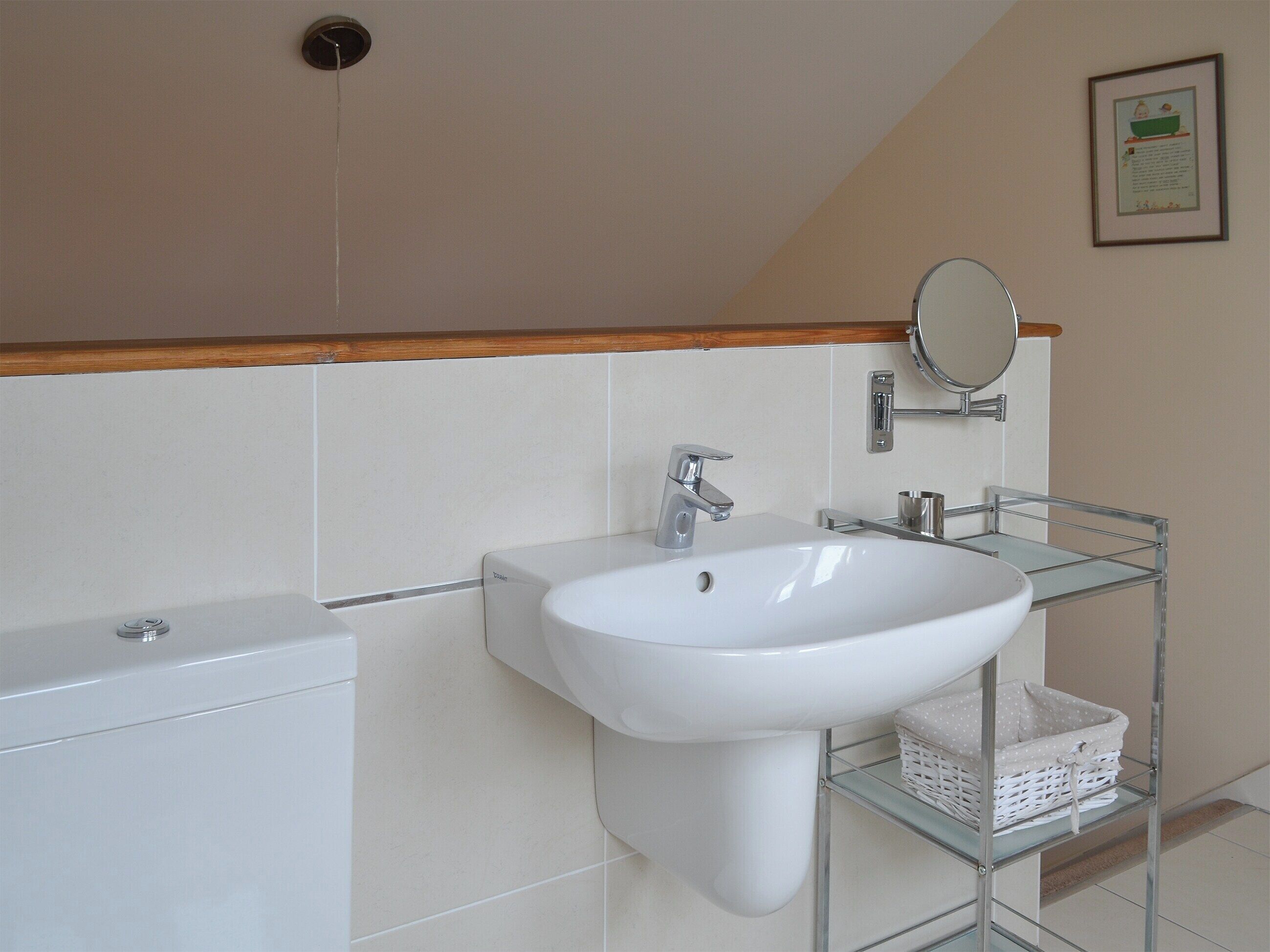 A bathroom with a white sink, a mirror, and a toilet. There is a shelf with a basket on it.