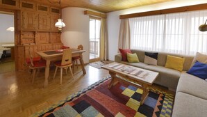 Una sala de estar acogedora con techo de madera, un sofá con cojines de colores, una mesa de comedor con sillas y un amplio ventanal con cortinas.