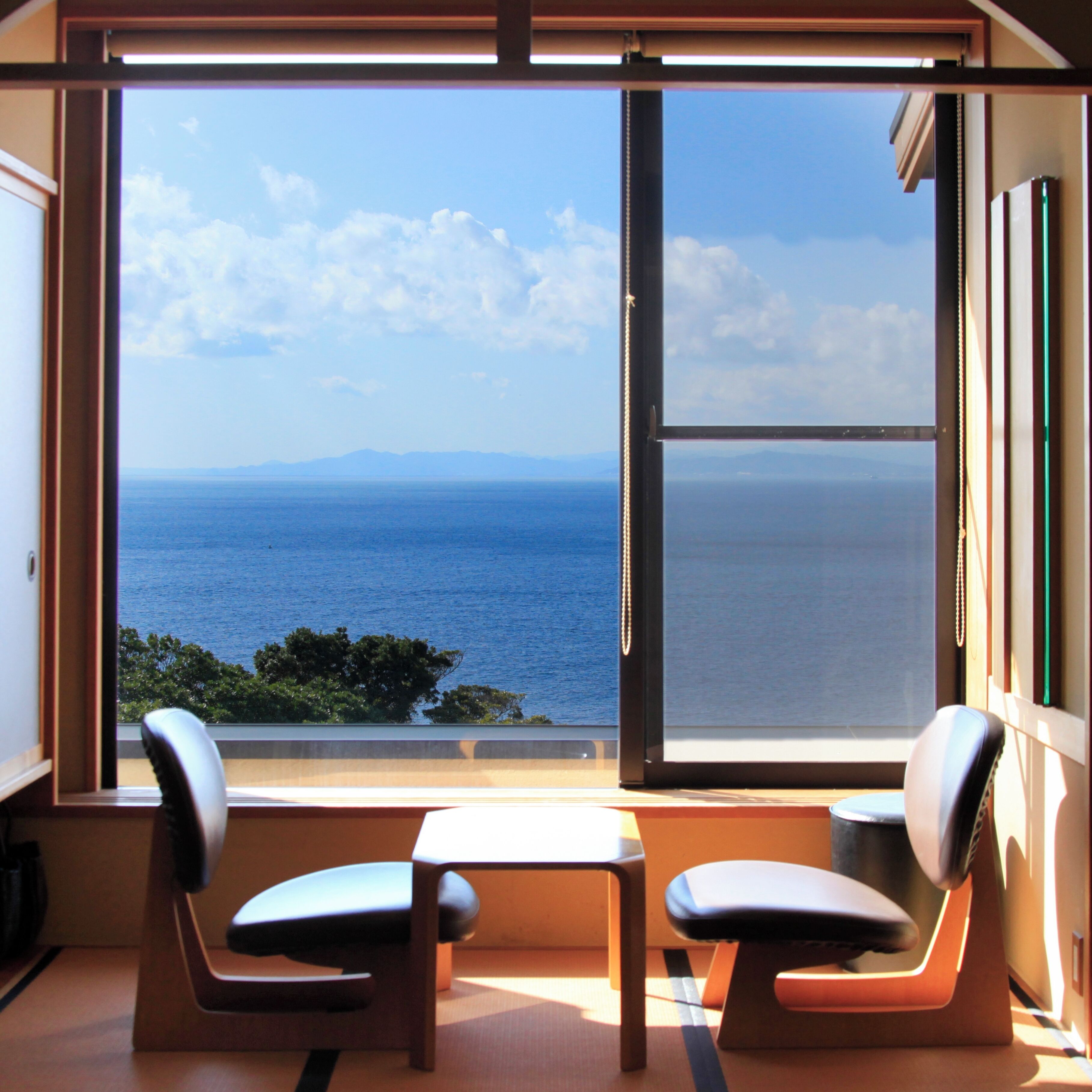 A room with a large window offering a view of the sea and distant mountains. There are two chairs and a small table in the room.