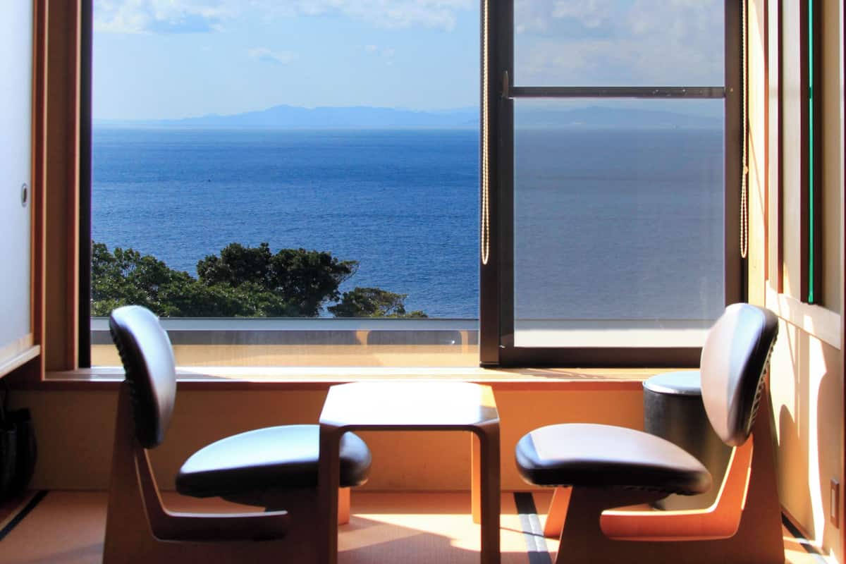 Quarto com uma grande janela que oferece vista para o mar e montanhas distantes. Há duas cadeiras e uma pequena mesa no quarto.