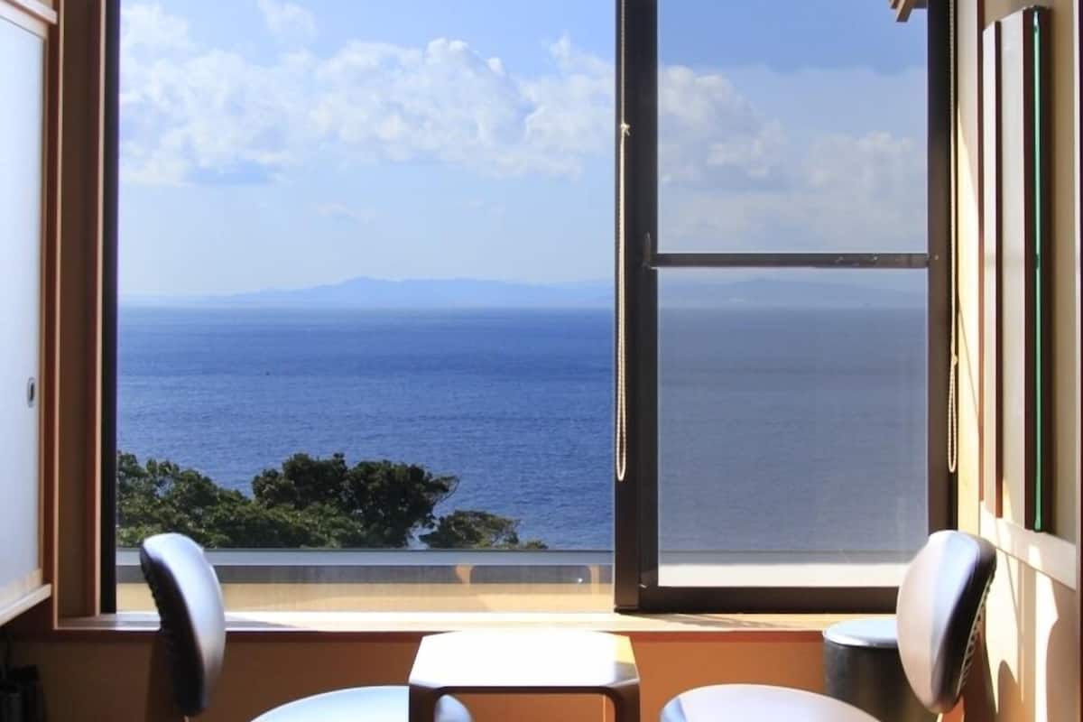 Quarto com uma grande janela com vista para o mar, duas cadeiras e uma mesa pequena.