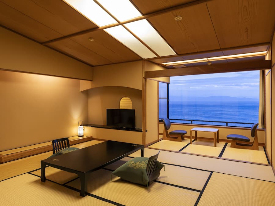 Ein Zimmer mit Tatami-Boden, einem niedrigen Tisch, Stühlen, einem Fernseher und Blick auf das Meer.
