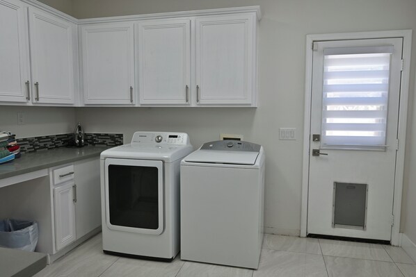 A laundry room with two white washing machines, a countertop with a coffee maker, and a door with blinds.