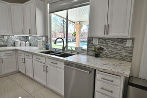 A kitchen with white cabinets, a stainless steel dishwasher, a double sink, and a view of a backyard with a pool.