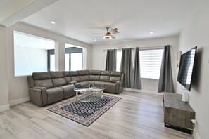 A modern living room with a sectional sofa, a glass coffee table, a ceiling fan, and a flat-screen TV mounted on the wall.